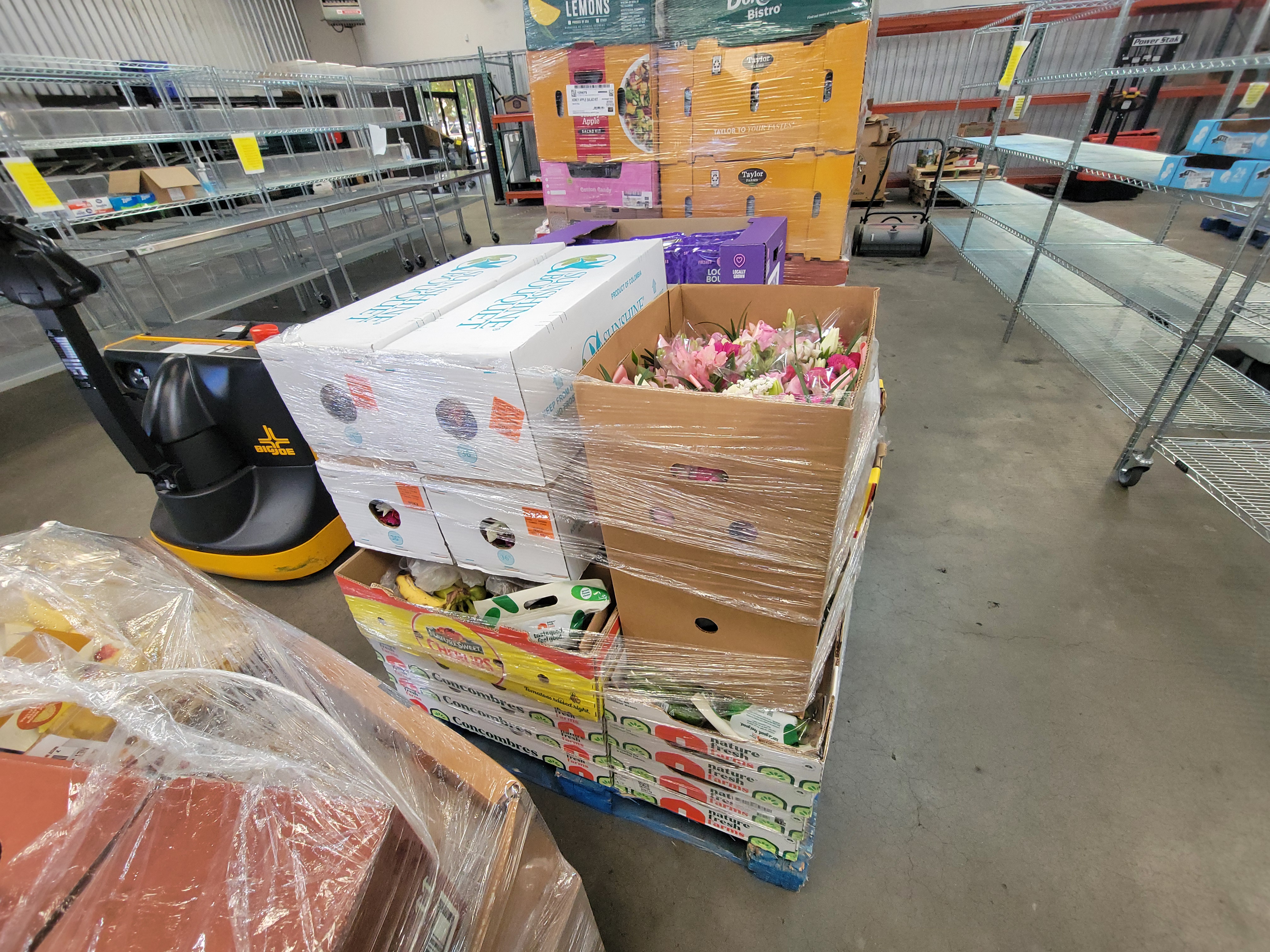 Boxes of flowers and produce on pallets wrapped in plastic wrap next to pallet jack.